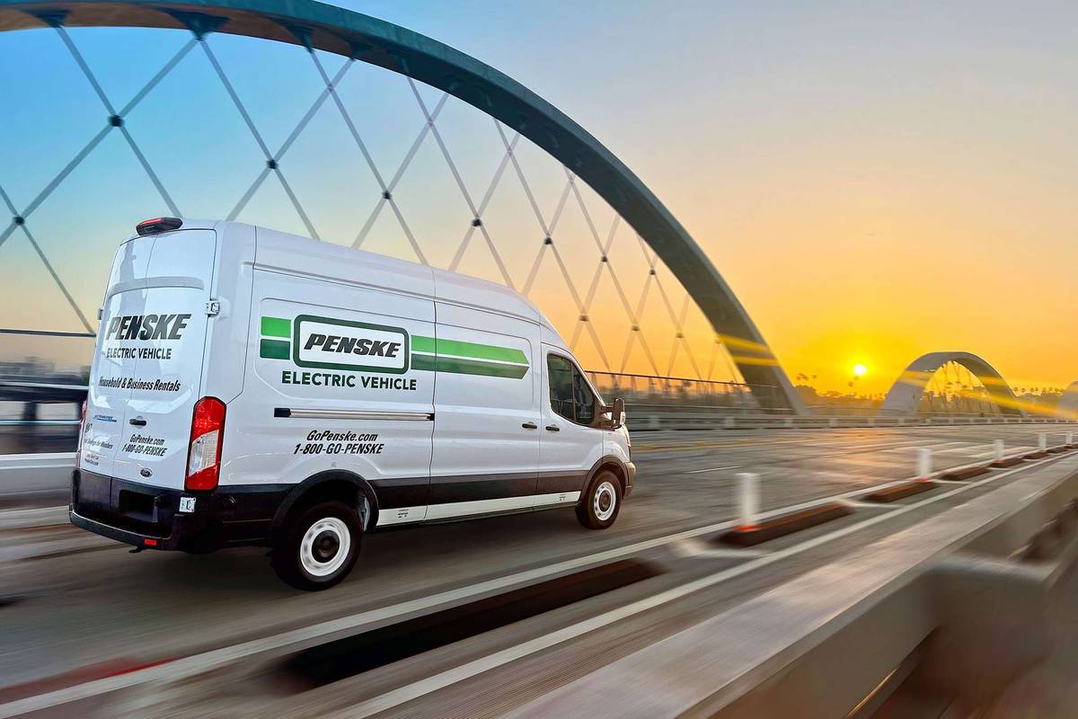 A white and green Penske electric cargo van drives over a bridge at sunset.