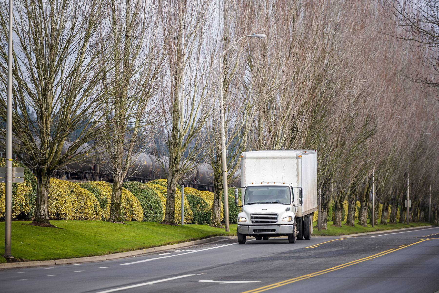 A white box truck drives up a street next to a train and green and yellow bushes.