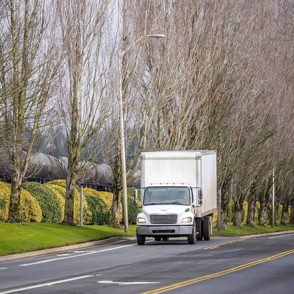 A white box truck drives up a street next to a train and green and yellow bushes.