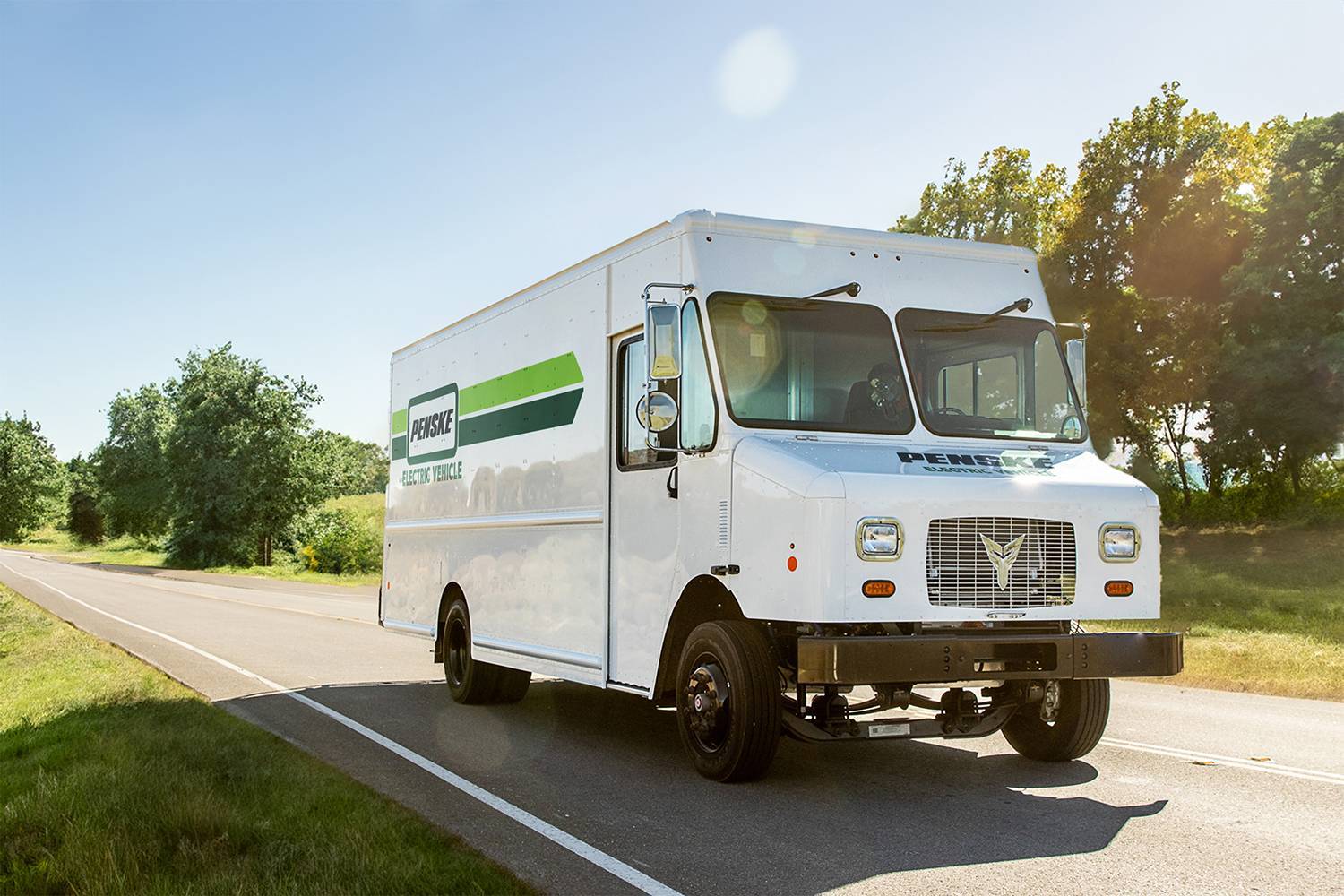 A white electric Penske step-van drives up the road on a sunny day.