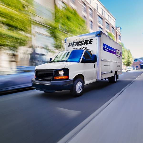 A white Penske box truck drives down city road.