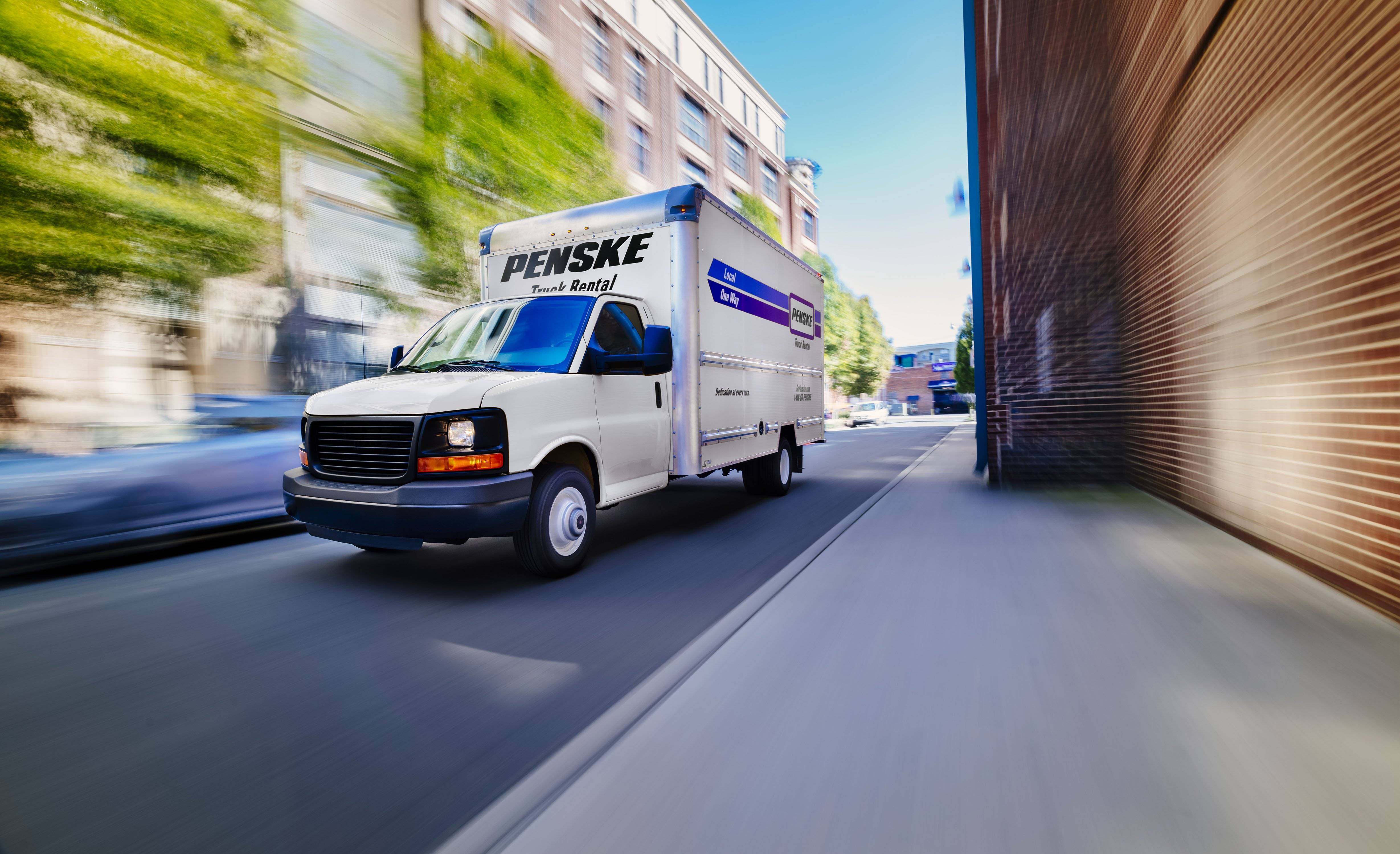 A white Penske box truck drives down city road.