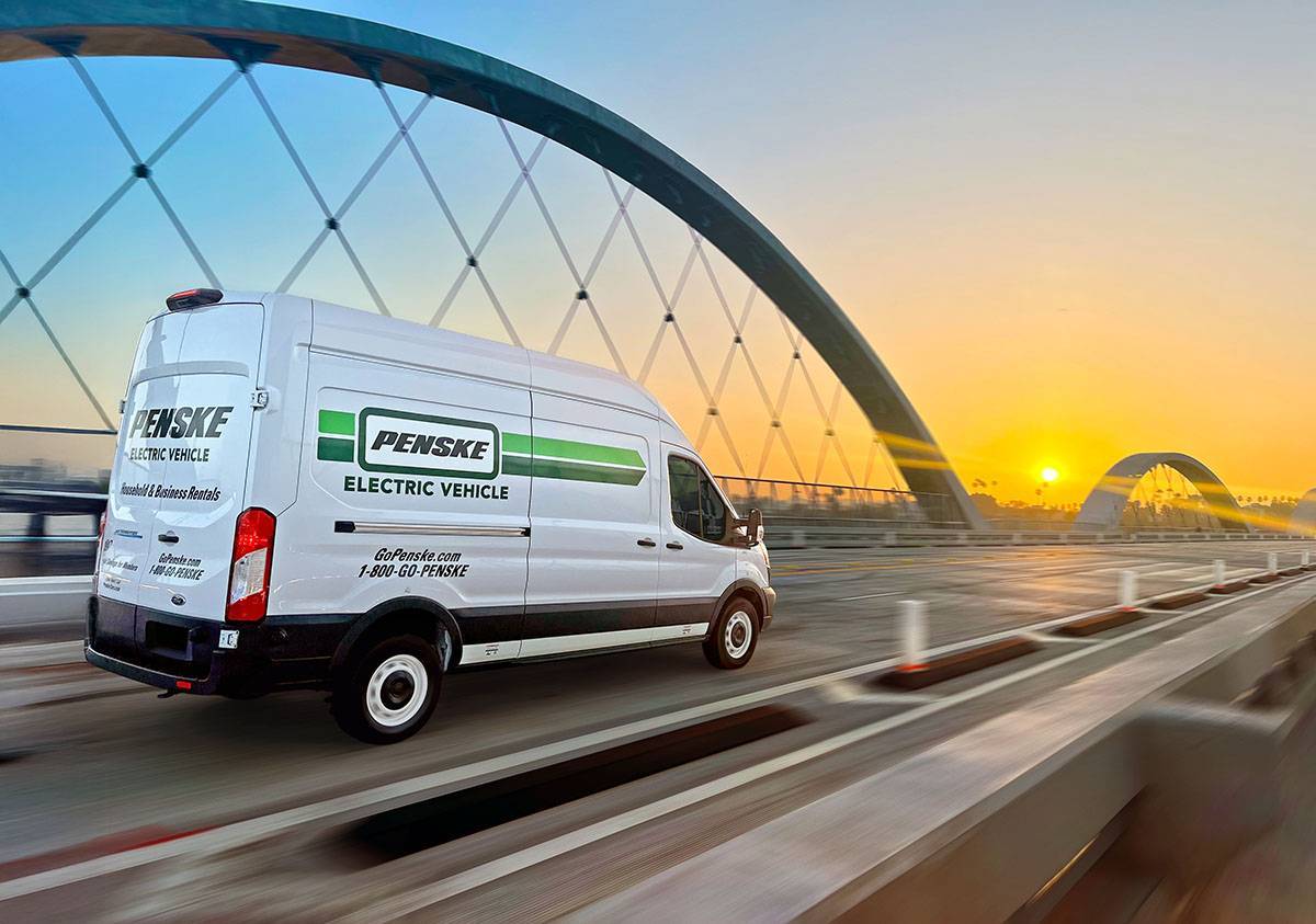 A white Penske electric cargo van drives on a freeway bridge at sunset.