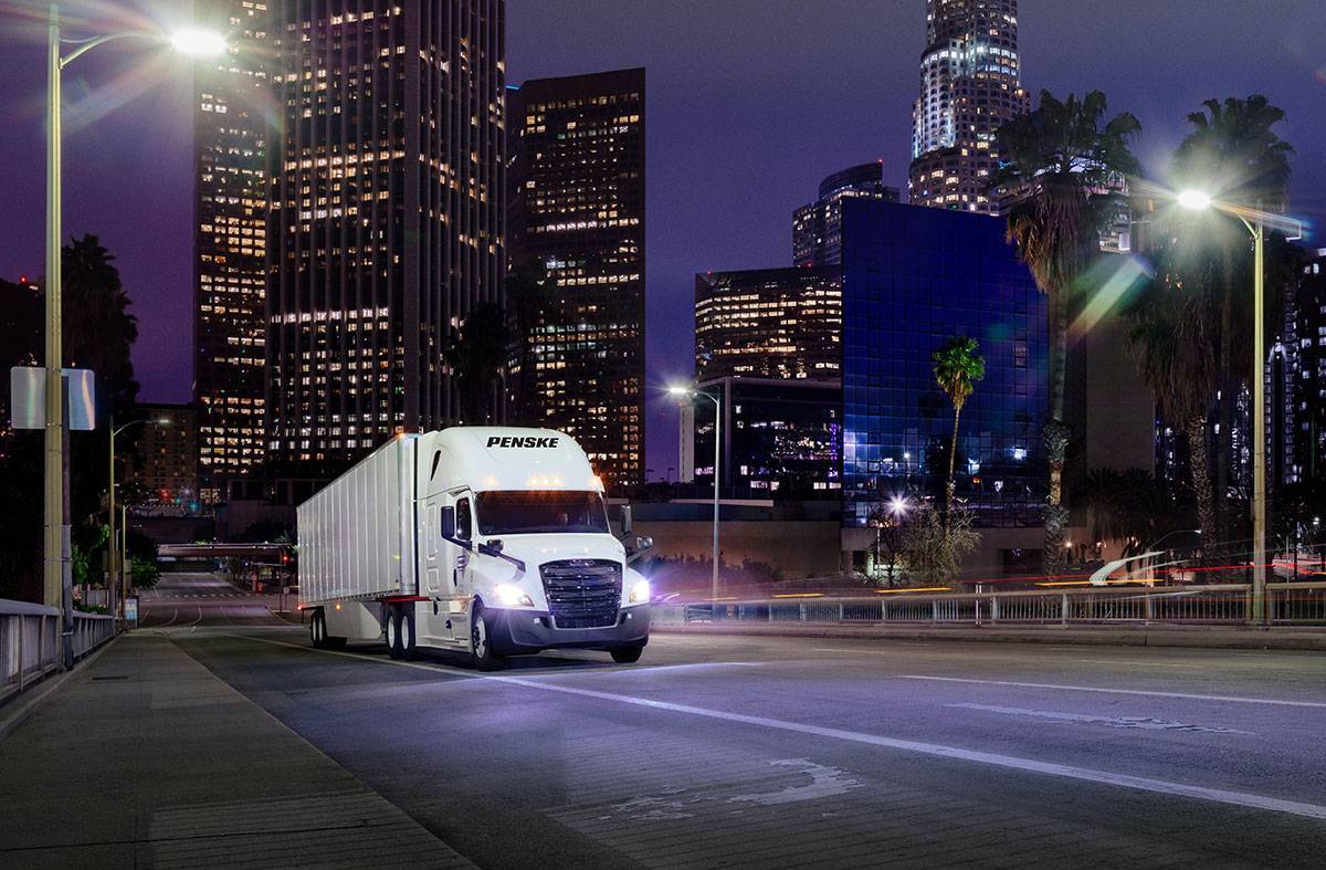 A white Penske semi truck at night driving in a dark city with skyscrapers and city lights.