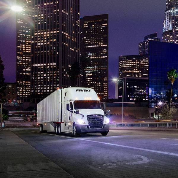 A white Penske semi truck at night driving in a dark city with skyscrapers and city lights.