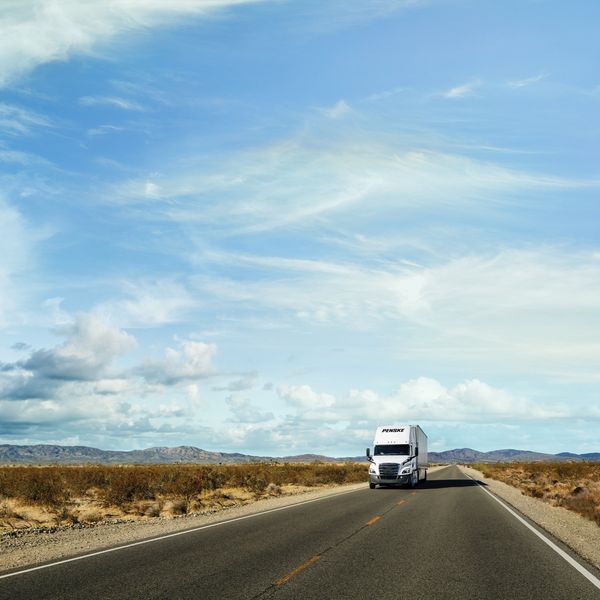 A white Penske semi-truck drives down a desert road. 