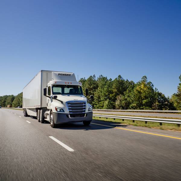 A white Penske semi-truck drives down a freeway next to blue skies and green trees.
