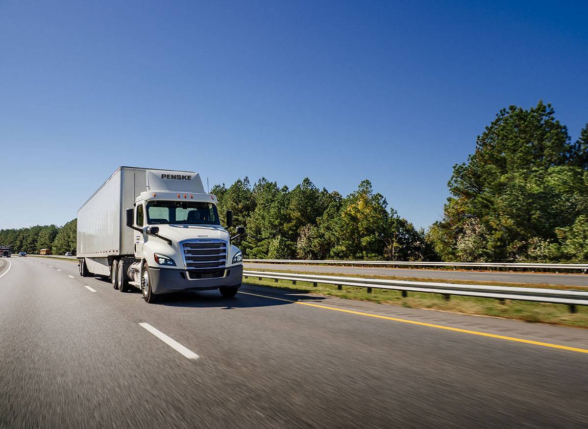 A white Penske semi-truck drives down a freeway surrounded by green trees.
