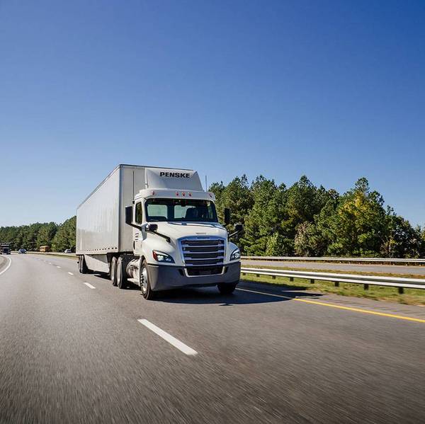 A white Penske semi-truck drives down a freeway surrounded by green trees.