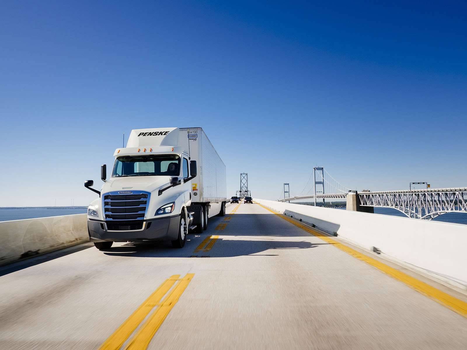 A white Penske semi-truck drives on a bridge over water.