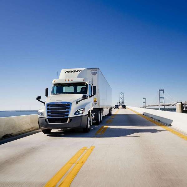 A white Penske semi-truck drives on a bridge over water.