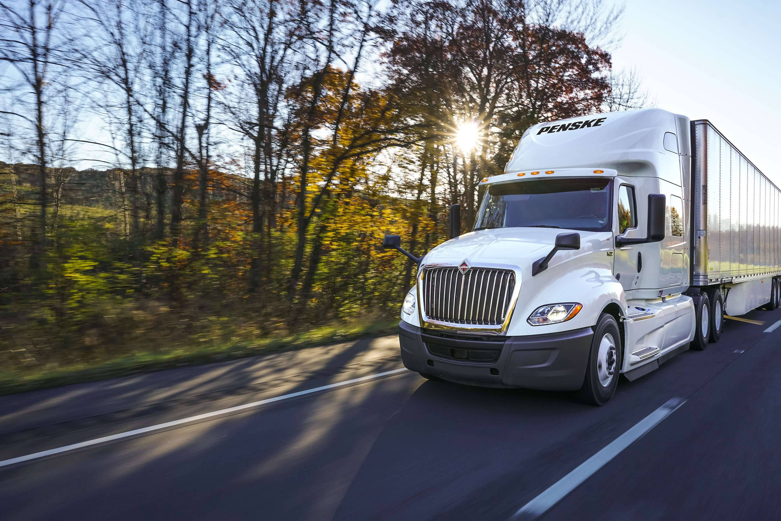 A white Penske semi-truck drives on a road past trees and the setting sun.