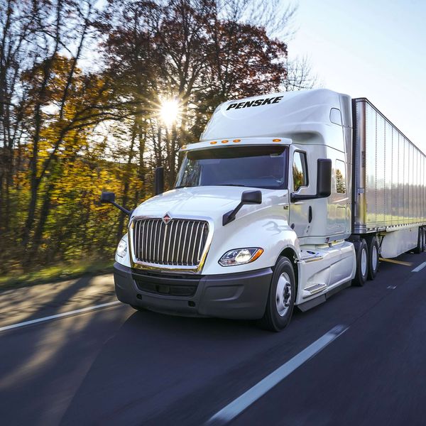 A white Penske semi-truck drives on a road past trees and the setting sun.