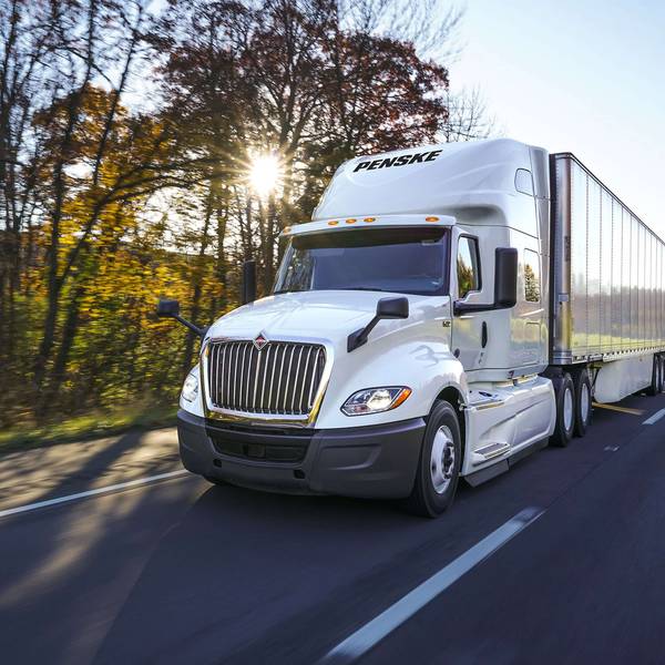 A white Penske semi-truck drives on a road past trees and the setting sun.