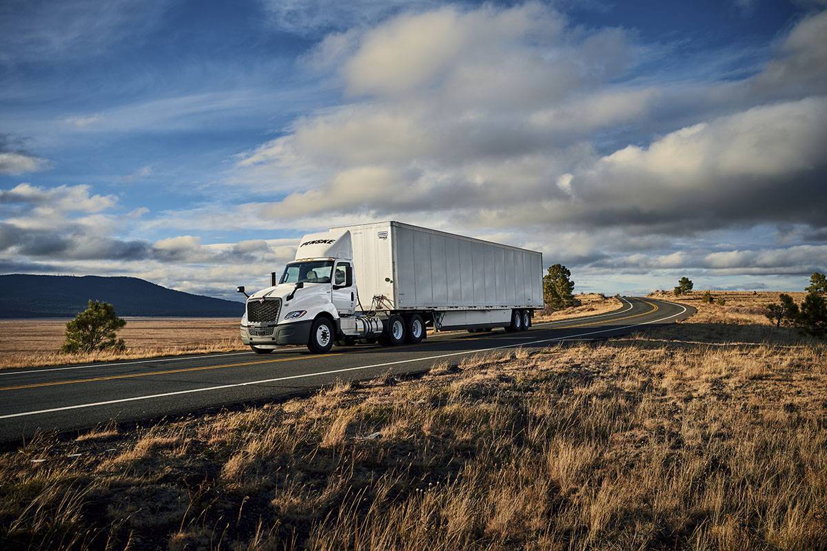 A white Penske semi-truck drives on a road surrounded by wheat fields under a blue sky.