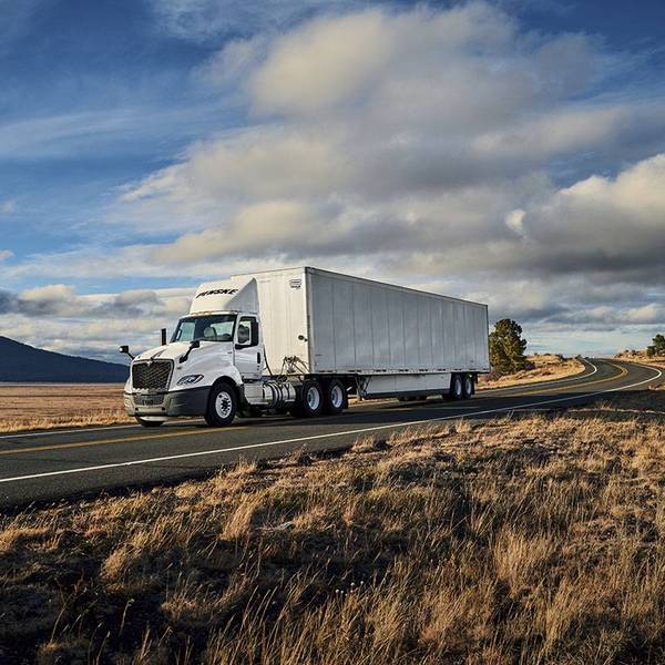 A white Penske semi-truck drives on a road surrounded by wheat fields under a blue sky.