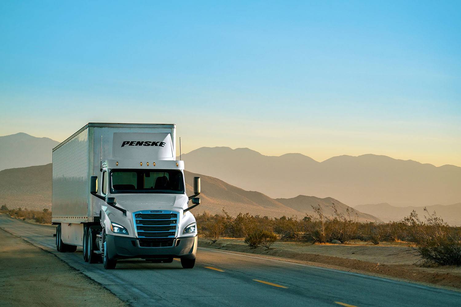 A white Penske semi-truck drives on a road with mountains in the background.