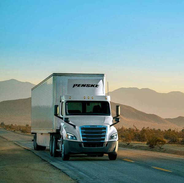 A white Penske semi-truck drives on a road with mountains in the background.