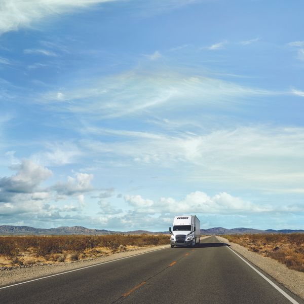 A white Penske semi-truck drives on an open desert road.