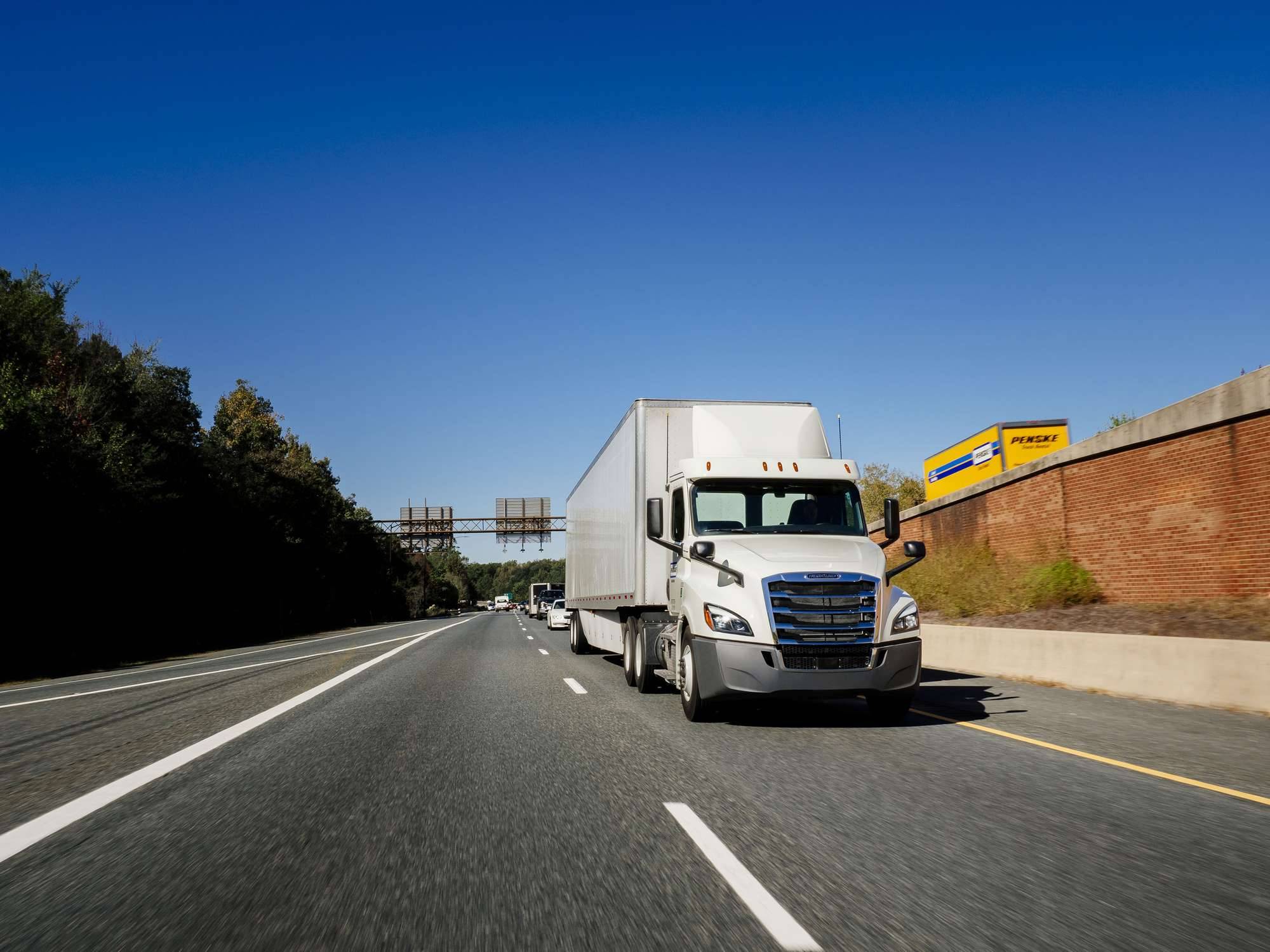 A white Penske semi-truck drives one way on a freeway past a yellow Penske box truck going the other way.