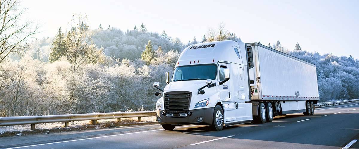 A white Penske semi-truck drives up a road in winter.