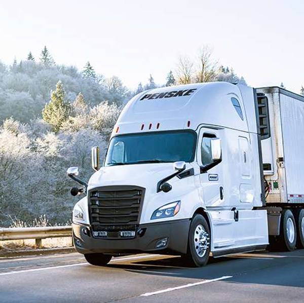 A white Penske semi-truck drives up a road in winter.