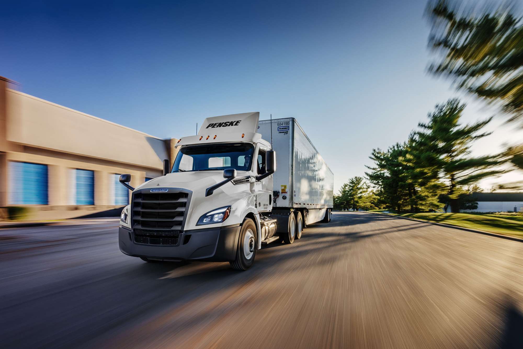 A white Penske semi-truck in motion on a street next to a building and trees.