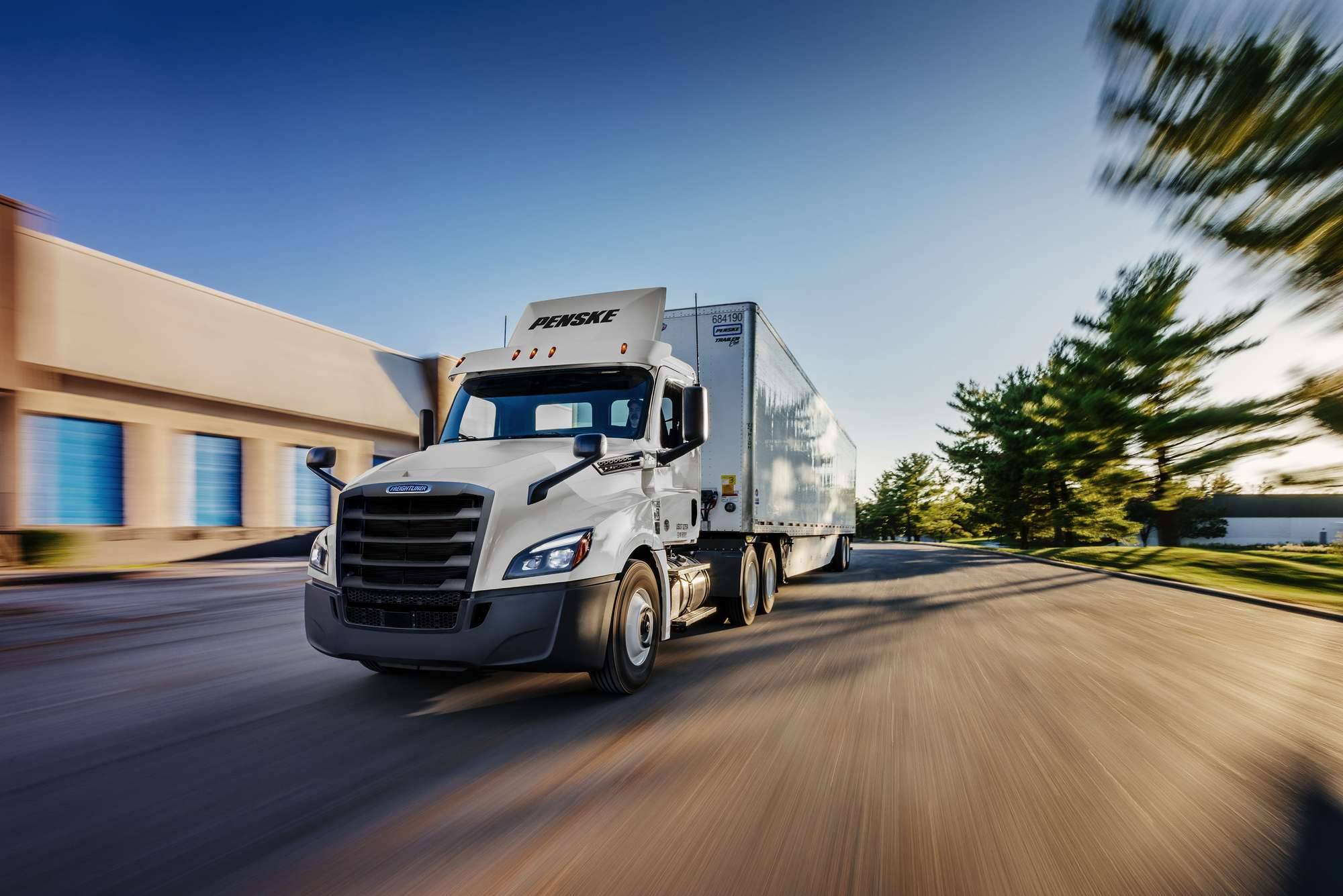 A white Penske semi-truck in motion on a street next to a building and trees.