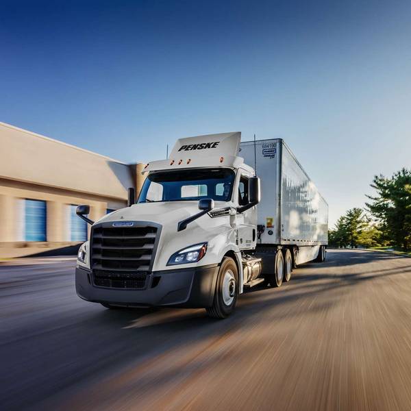A white Penske semi-truck in motion on a street next to a building and trees.