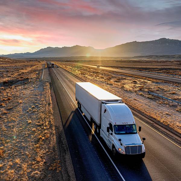 A white semi-truck and trailer drives up a desert road at dusk.