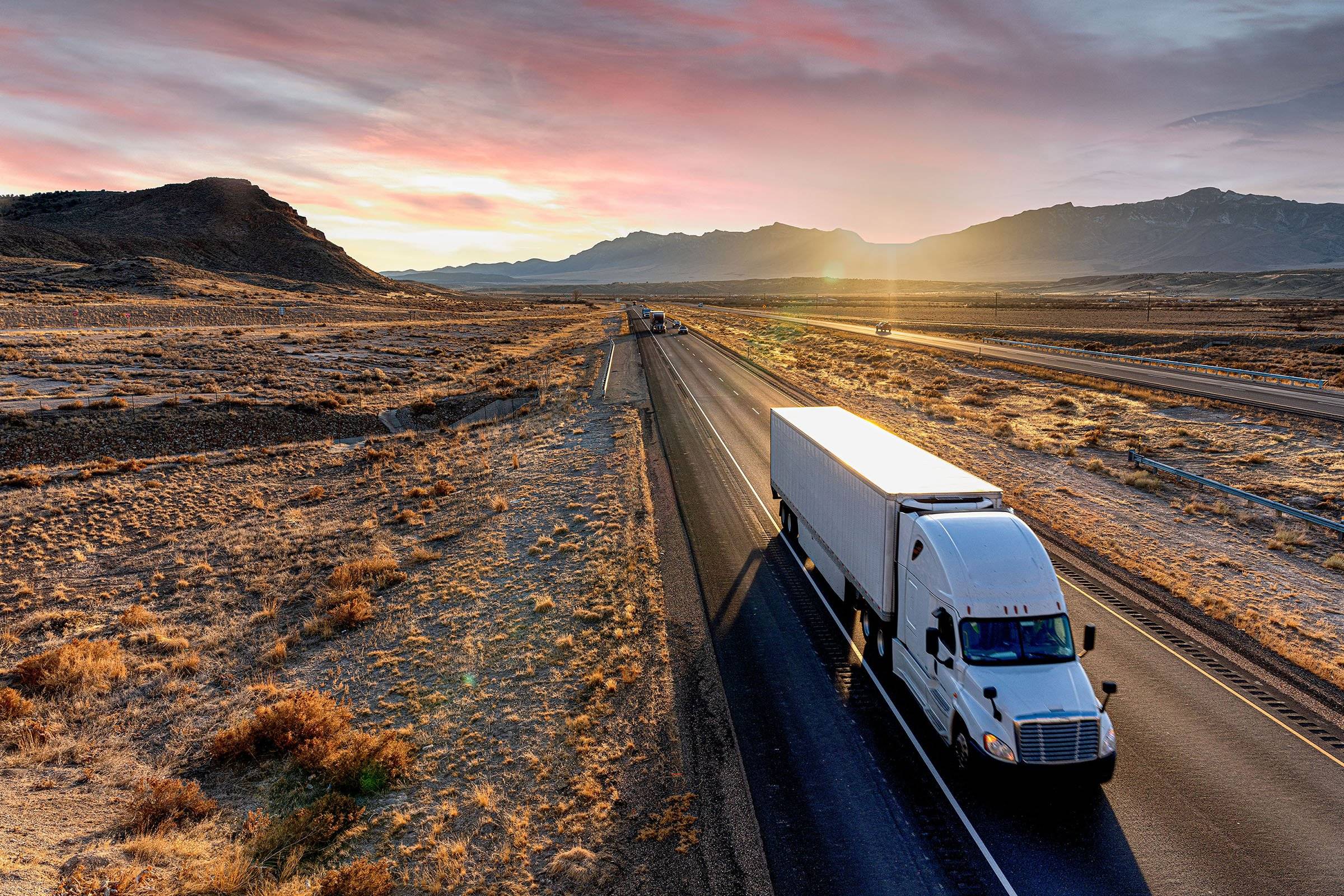 A white semi-truck and trailer drives up a desert road at dusk.
