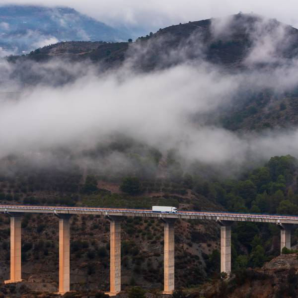 A white semi-truck drives across a bridge through foggy mountains.