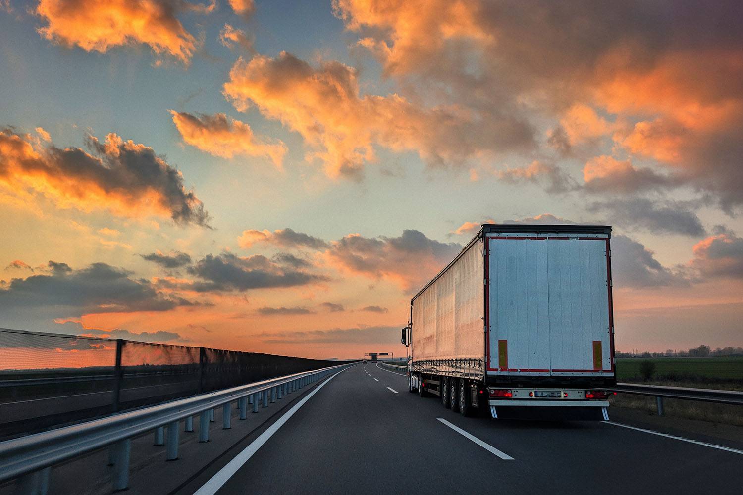 A white semi-truck drives away from the camera under an orange sunset sky.
