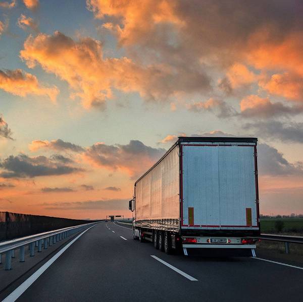 A white semi-truck drives away from the camera under an orange sunset sky.