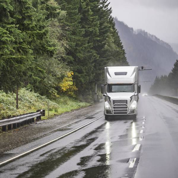 A white semi truck drives on a rainy road surrounded by green trees.