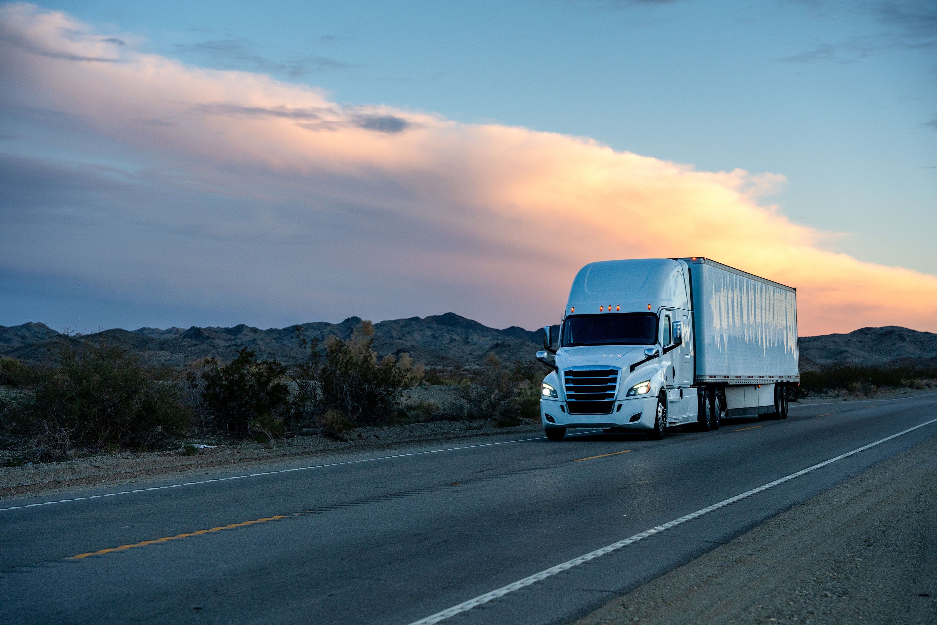 A white semi-truck drives up a road at sunset.