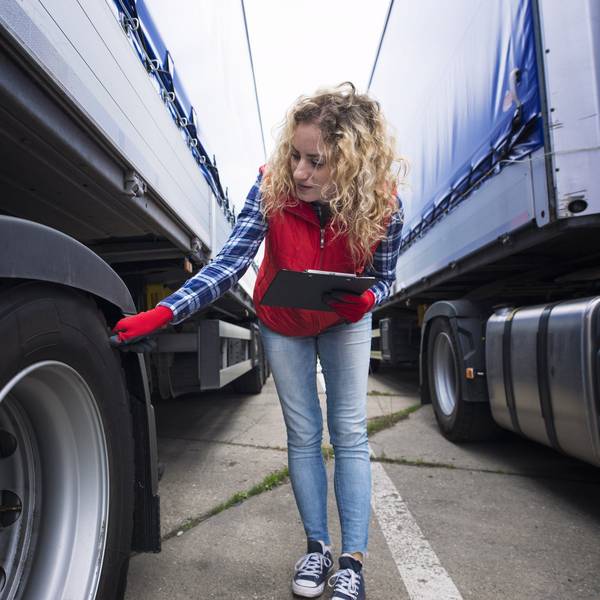 A woman holds a clipboard and checks the tire of a semi-truck.