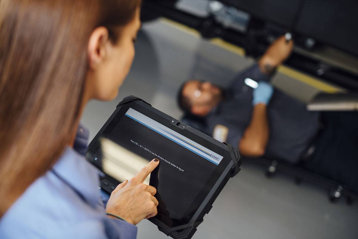 A woman touches an iPad while a man works on the bottom of a vehicle in the background.
