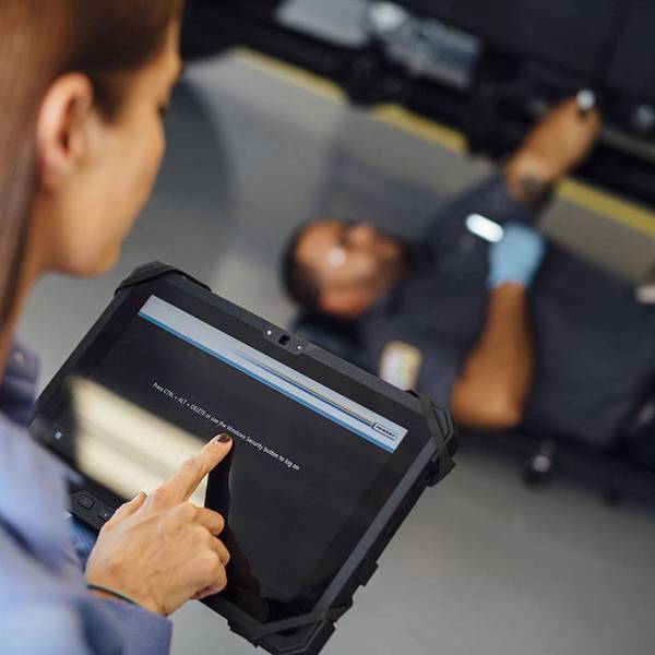 A woman touches an iPad while a man works on the bottom of a vehicle in the background.