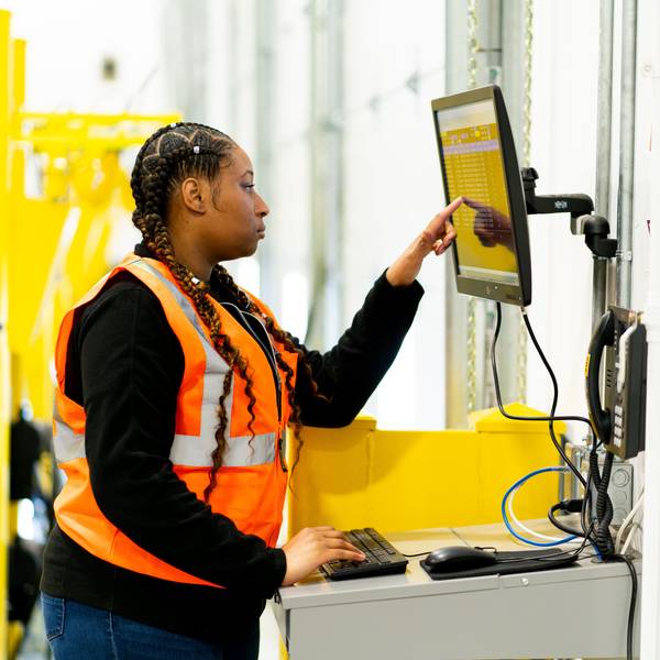 A woman wearing a safety vest works on a computer while in a warehouse.