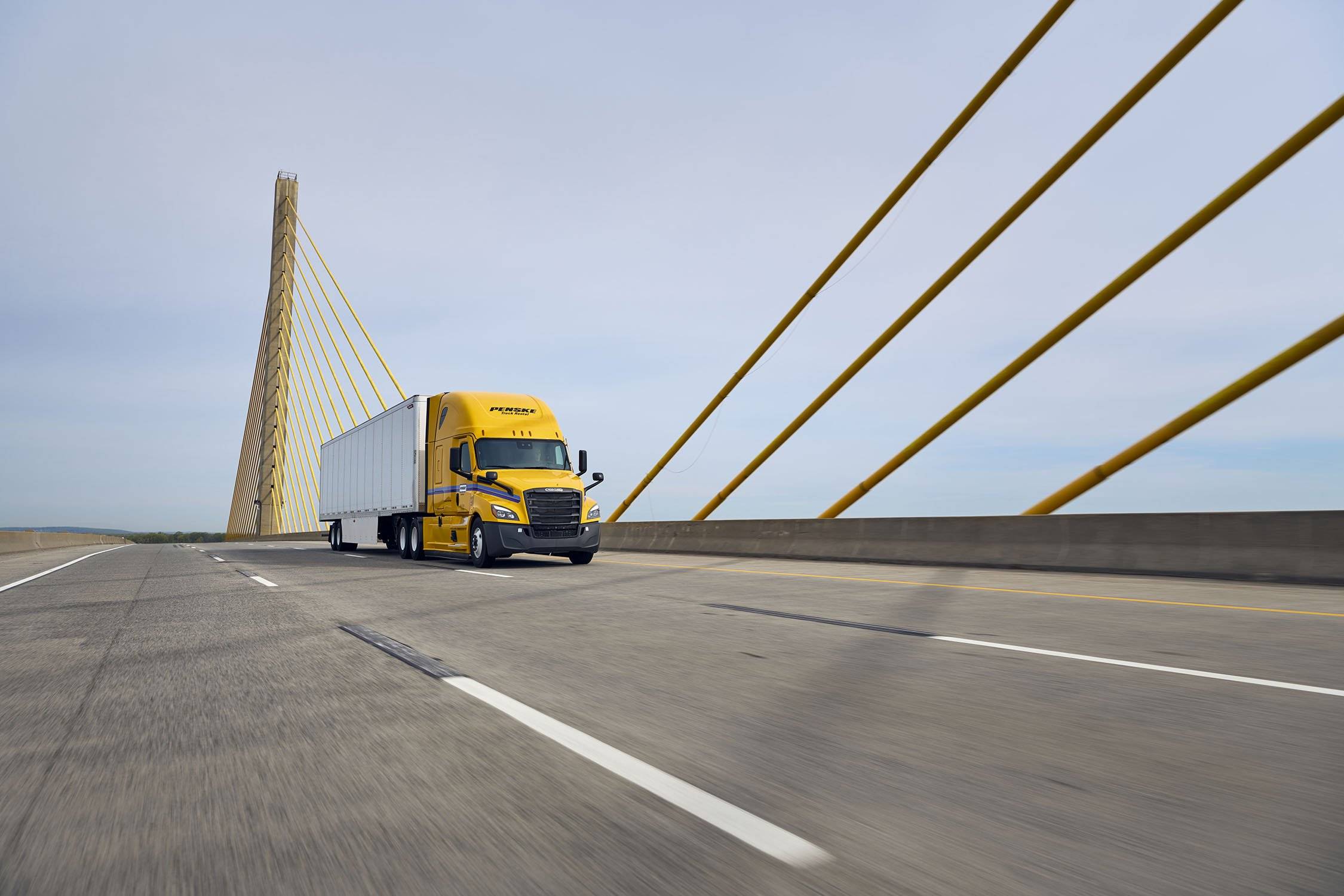 A yellow and white Penske semi-truck drives across a bridge.