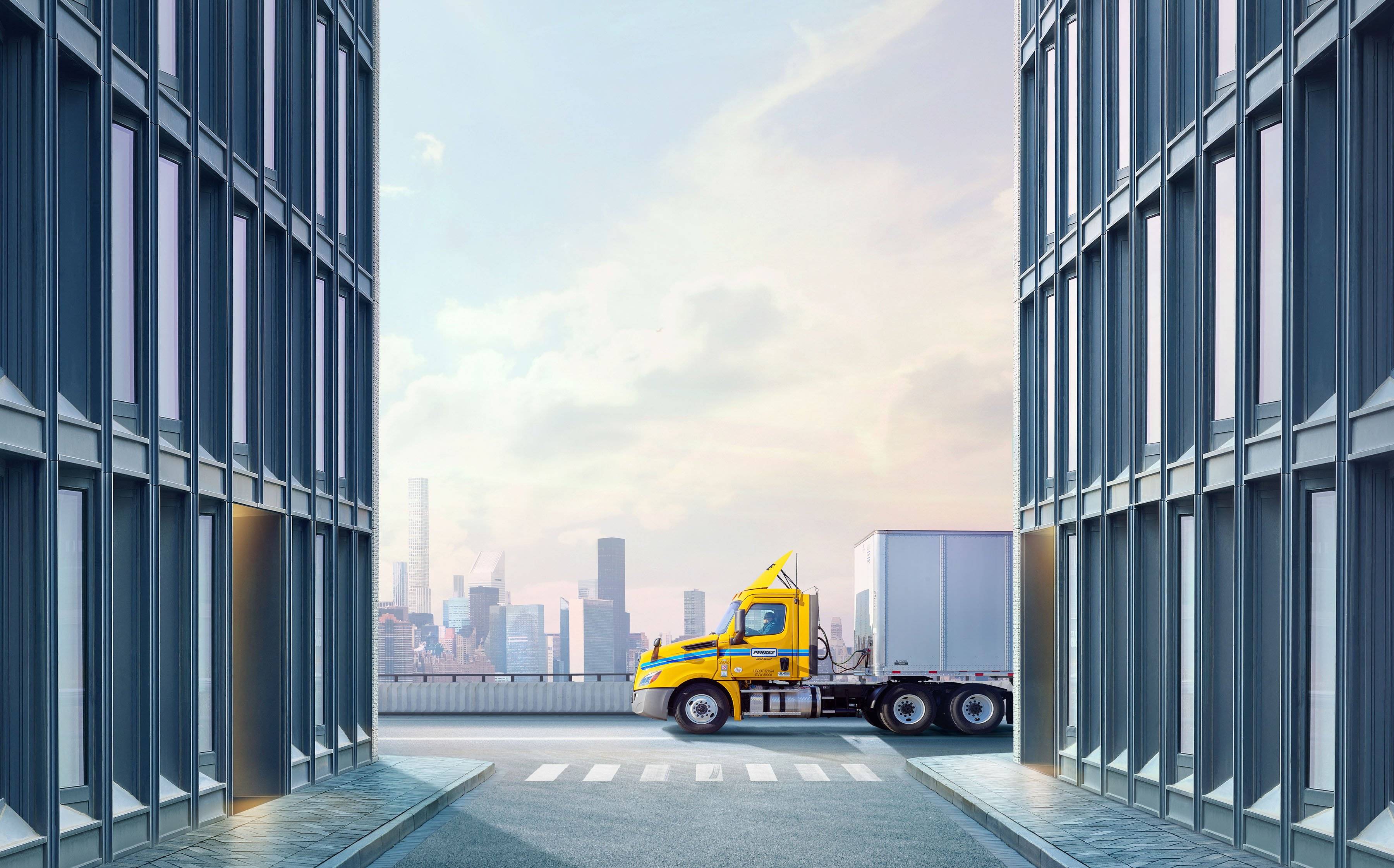 A yellow and white Penske semi-truck drives along a city road.