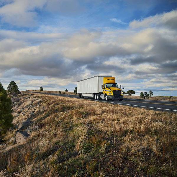 A yellow and white Penske semi-truck drives along a country road under a blue cloudy sky.