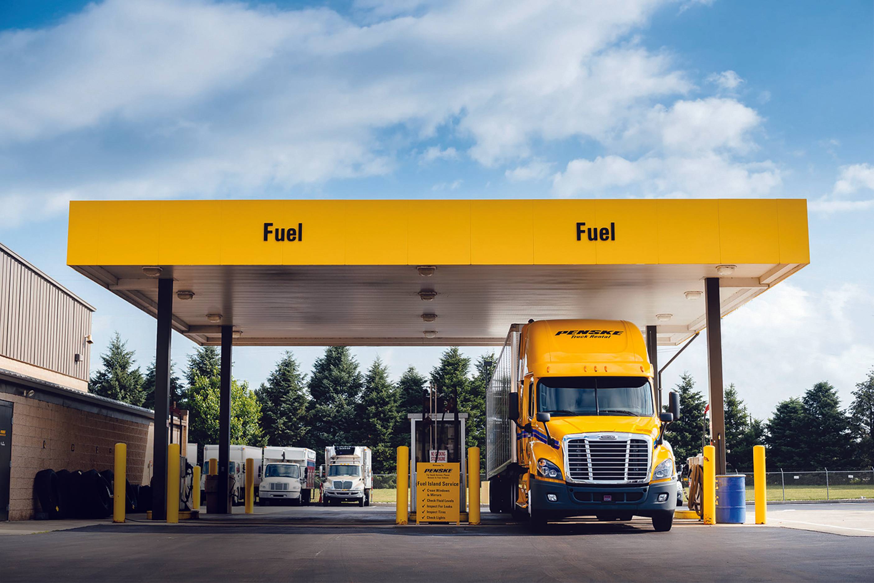 A yellow Penske semi-truck pulled up at a fuel station with yellow Fuel banner overtop.