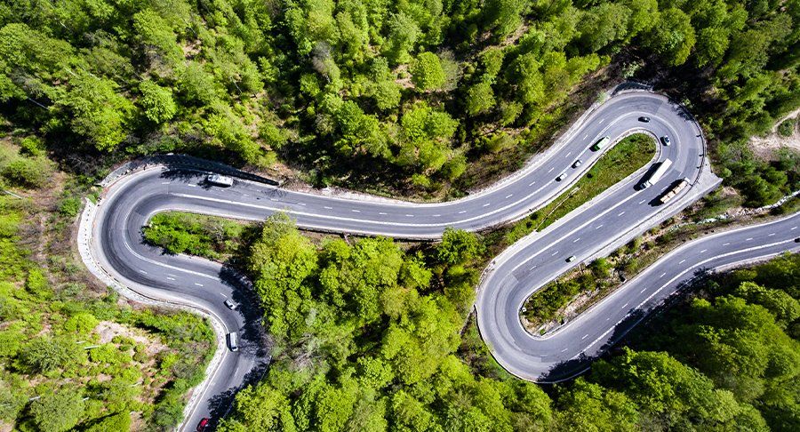 An aerial shot of trucks driving on a curvy road with lots of green trees.