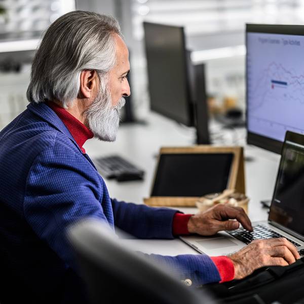 An older man sits at a desk in front of three computer screens.