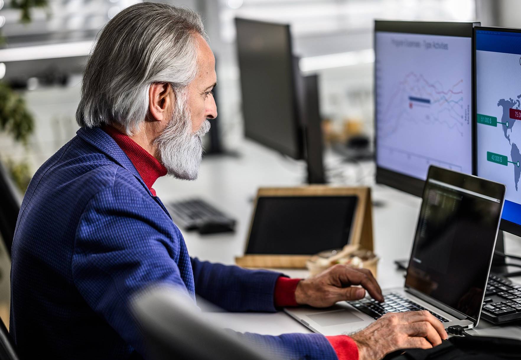 An older man sits at a desk in front of three computer screens.