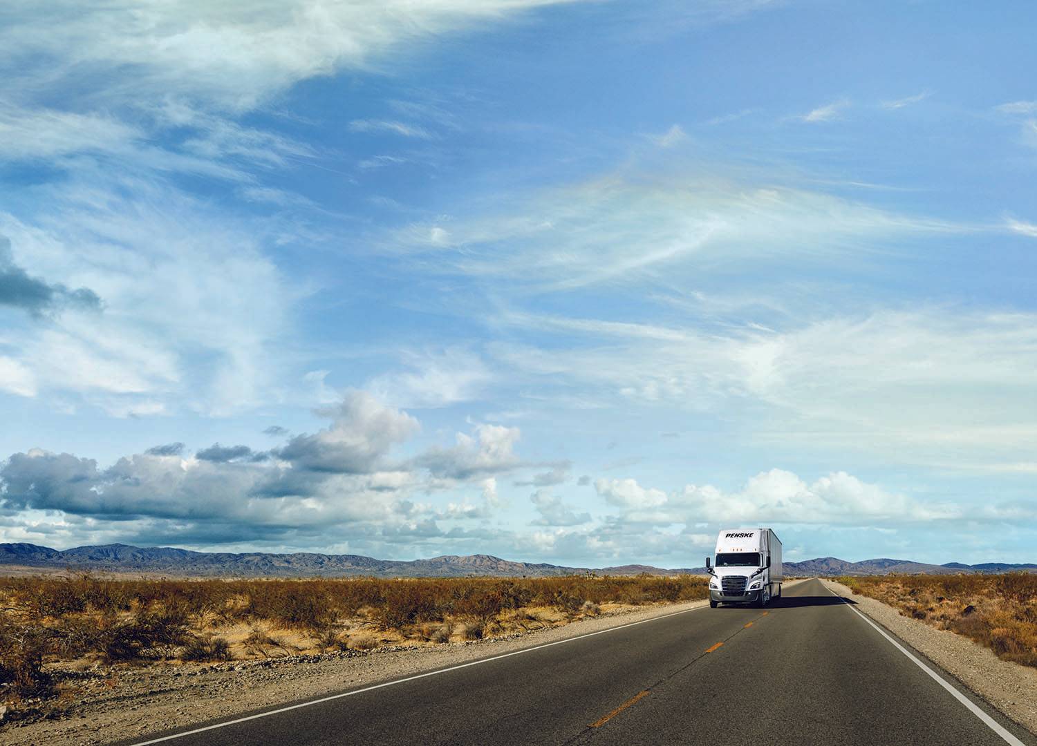 Blue cloudy sky, desert landscape with a white Penske semi-truck driving on a road.