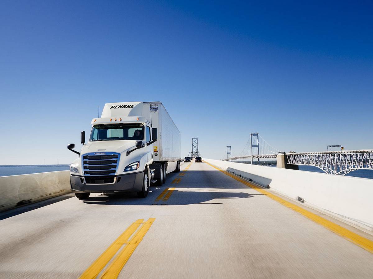 Blue sky with a white Penske semi-truck driving on a highway bridge.