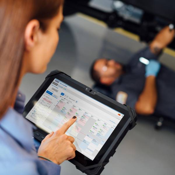 Mechanic under car, person using tablet for scheduling in foreground.